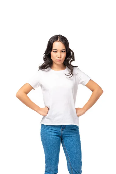 Mujer asiática enojada en camiseta blanca y jeans azules tomados de las manos en las caderas aisladas en blanco - foto de stock