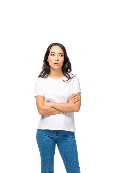 Mujer asiática reflexiva en camiseta blanca y jeans azules de pie con brazos cruzados aislados en blanco - foto de stock