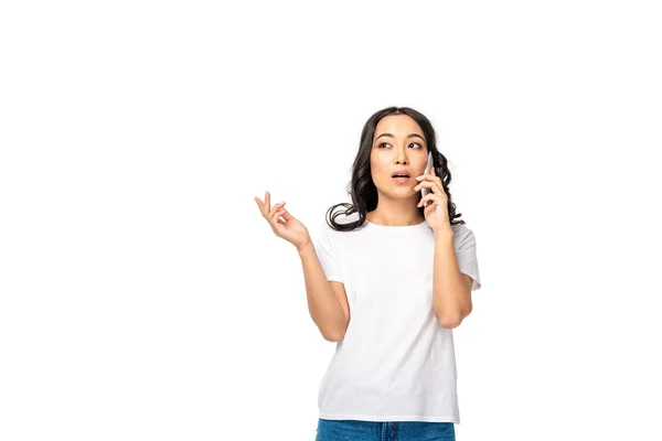 Задумчивая азиатская девушка в белой футболке и синих джинсах разговаривает на смартфоне изолированы на белом — стоковое фото