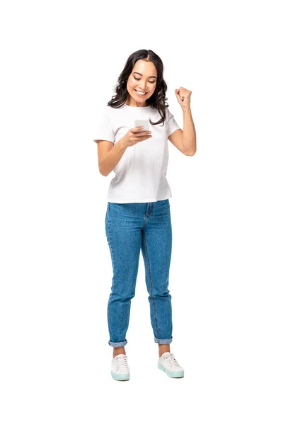 Smiling happy asian girl in white t-shirt and blue jeans using smartphone and raising fist isolated on white — Stock Photo