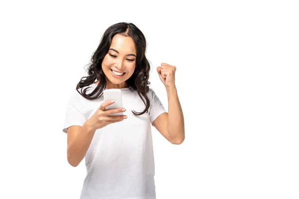 Sonriente joven asiático mujer en blanco camiseta usando smartphone y levantando puño aislado en blanco - foto de stock