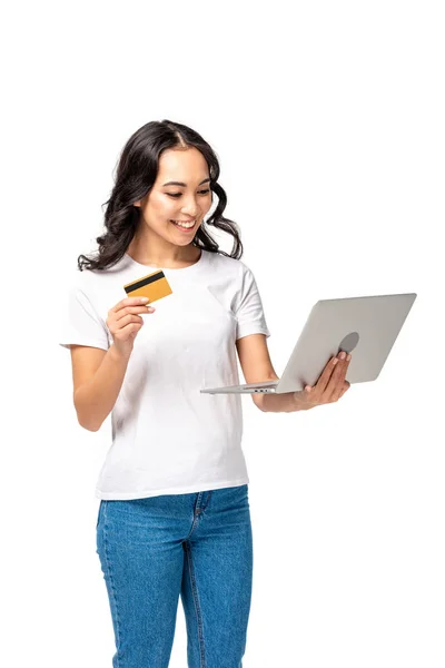 Усміхнена азіатська жінка в білій футболці і синіх джинсах використовує ноутбук і тримає кредитну картку ізольовано на білому — стокове фото