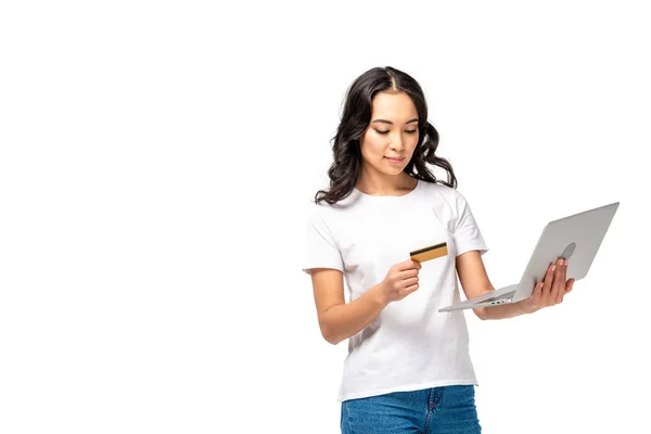 Konzentrierte Asiatin in weißem T-Shirt und blauer Jeans mit Laptop und Kreditkarte isoliert auf weißem Hintergrund — Stockfoto