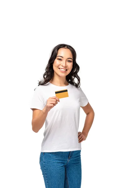Belle souriante asiatique femme montrant carte de crédit et tenant la main sur la hanche isolé sur blanc — Photo de stock