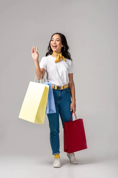 Moda vestida joven mujer asiática sosteniendo coloridas bolsas de compras y saludando con la mano levantada en gris - foto de stock