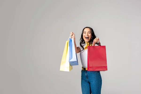 Smiling asian woman holding colorful shopping bags on raised hands and looking at camera isolated on grey — Stock Photo
