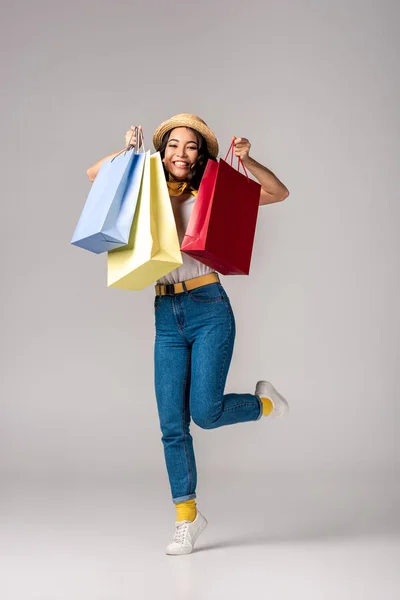 Feliz mujer asiática vestida de moda sosteniendo coloridas bolsas de compras en la mano levantada en gris - foto de stock