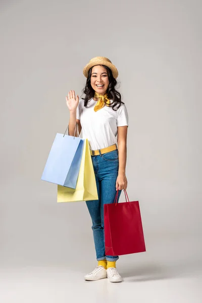 Lächelnde glückliche asiatische Frau mit trendigem Hut, bunte Einkaufstüten in der Hand und winkende Hand auf grau — Stockfoto