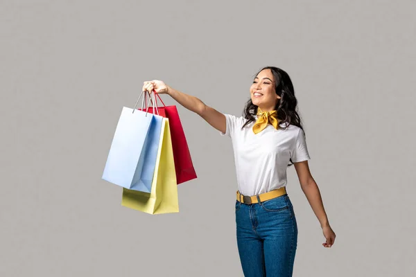 Happy smiling asian girl holding colorful shopping bags in one stretched hand isolated on grey — Stock Photo