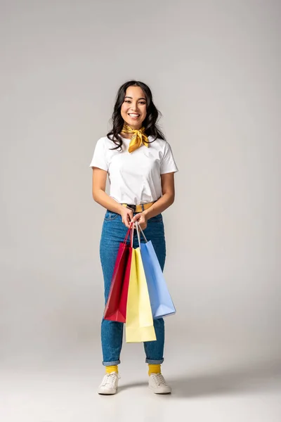 Приваблива усміхнена азіатка тримає барвисті сумки для покупок на сірому — стокове фото