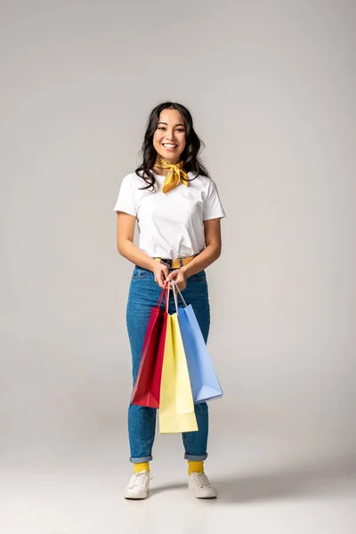 Attraente sorridente donna asiatica che tiene borse colorate su grigio — Foto stock