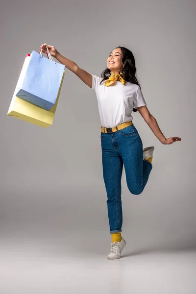 Mujer asiática feliz sosteniendo coloridas bolsas de compras en mano estirada en gris - foto de stock