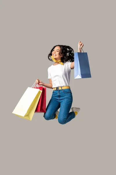 Захоплена азіатська жінка тримає барвисті сумки для покупок і щасливо стрибає однією піднятою рукою ізольовано на сірому — стокове фото
