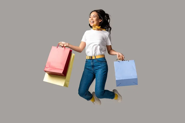 Rindo asiático mulher segurando colorido sacos de compras e felizmente pulando isolado no cinza — Fotografia de Stock