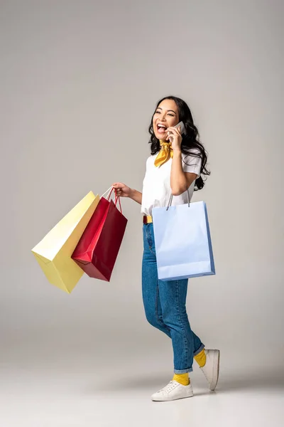 Sonriente joven asiática mujer sosteniendo coloridas bolsas de compras y hablando en el teléfono inteligente en gris - foto de stock