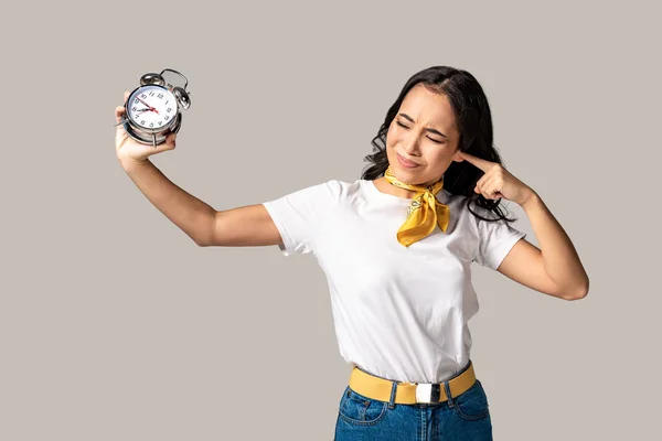 Asian woman with closed eyes holding alarm clock in stretched hand and closing ear with forefinger isolated on grey — Stock Photo