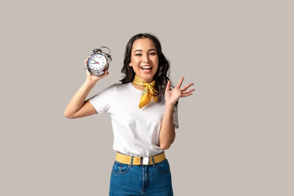 Mujer asiática feliz en camiseta blanca y vaqueros azules sosteniendo reloj despertador y agitando la mano aislada en gris - foto de stock