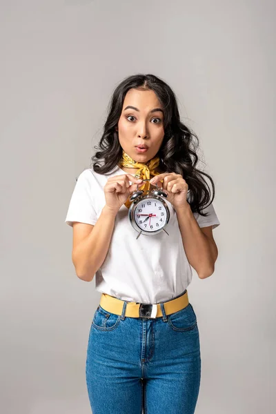 Surprised young asian woman holding alarm clock in front of herself isolated in grey — Stock Photo