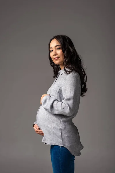Jolie jeune femme enceinte asiatique hoding ventre avec les mains isolées sur gris — Photo de stock