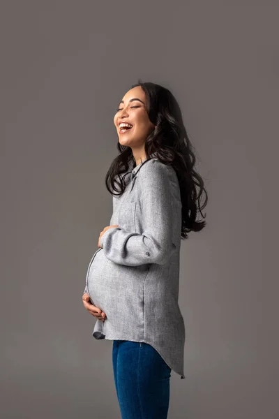 Счастливая беременная азиатка держит животик руками и смеется изолированно на серой — стоковое фото