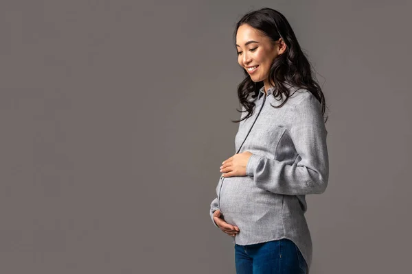 Smiling pregnant asian woman holding tummy with hands isolated on grey — Stock Photo