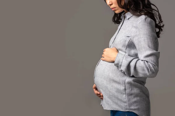 Vista parcial da mulher asiática grávida grave segurando a barriga com as mãos isoladas no cinza — Fotografia de Stock