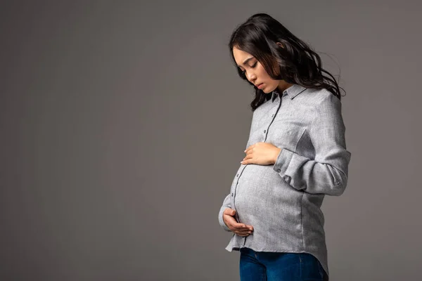 Серьёзно беременная азиатка в серой рубашке и синих джинсах, смотрящая на желудок, изолированный на сером — стоковое фото