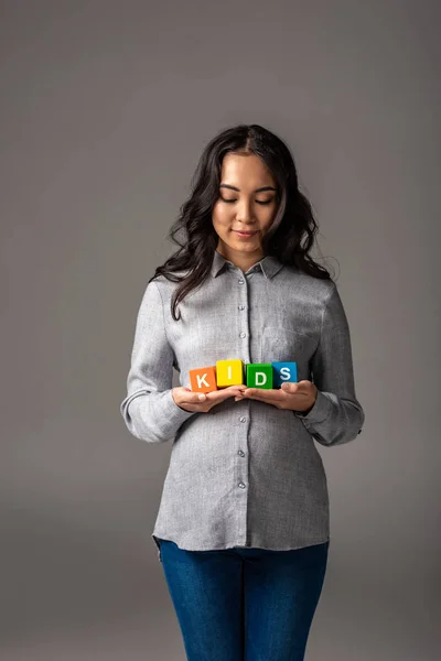 Smiling pregnant young asian woman holding alphabet cubes with word kids isolated on grey — Stock Photo