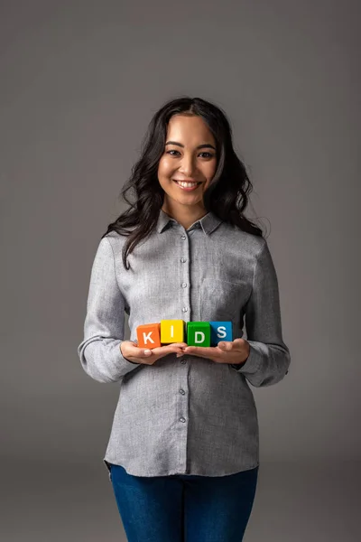 Cheerful pregnant young asian woman holding alphabet cubes with word kids on grey — Stock Photo