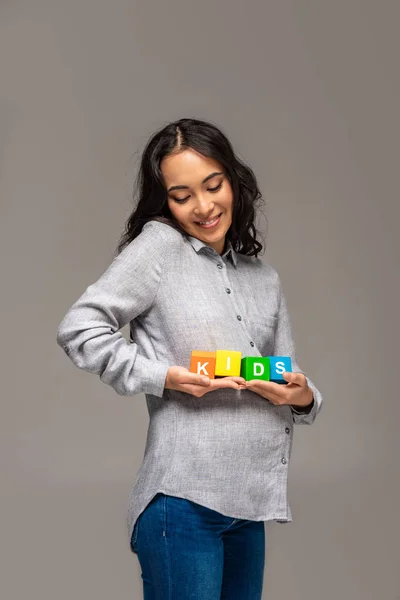 Улыбающаяся беременная азиатка с кубиками алфавита и детскими буквами, изолированными на сером — стоковое фото