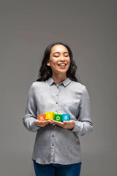 Lächelnd schwangere asiatische junge Frau hält bunte Buchstabenwürfel mit Wortkindern auf grau isoliert — Stockfoto