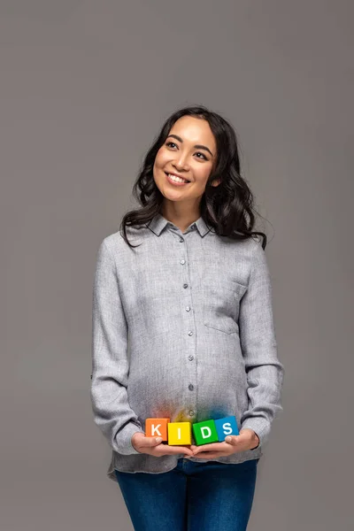 Sonriendo embarazada asiática mujer sosteniendo alfabeto cubos con palabra niños aislado en gris - foto de stock