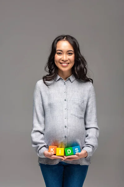 Smiling pregnant young asian woman holding alphabet cubes with word kids isolated on grey — Stock Photo