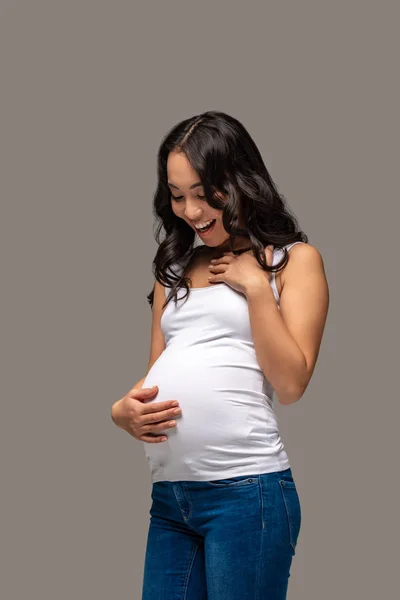 Улыбающаяся беременная азиатка в белой футболке и синих джинсах, смотрящая на живот, изолированный на сером — стоковое фото