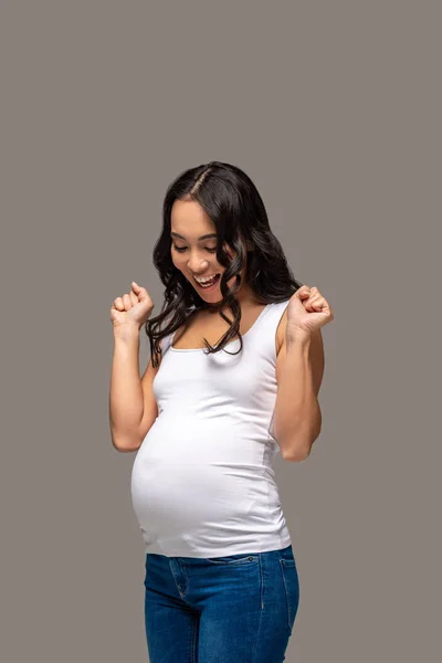 Felice donna asiatica incinta in t-shirt bianca guardando pancia e alzando le mani isolate sul grigio — Foto stock