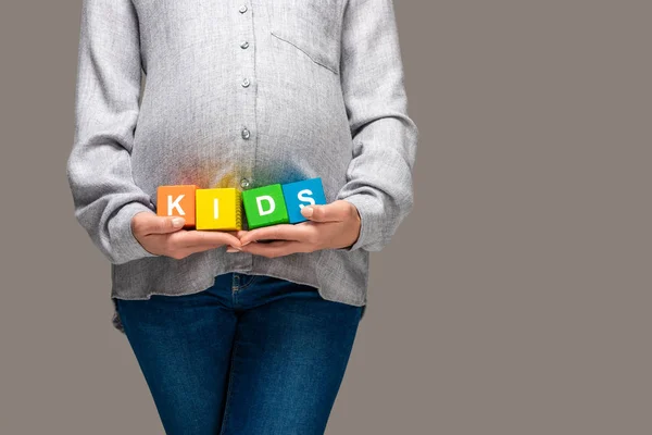 Vista parcial de la mujer asiática embarazada en camisa gris y jeans azules sosteniendo cubos de alfabeto con palabra niños debajo del vientre aislado en gris - foto de stock