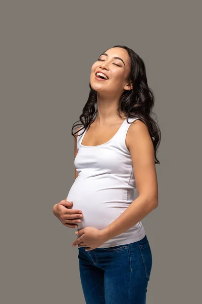 Feliz embarazada asiática con los ojos cerrados en camiseta blanca y vaqueros azules sosteniendo el vientre con las manos y riendo aislado en gris — Stock Photo