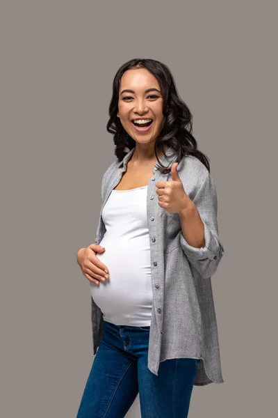 Sorrindo grávida asiático mulher tocando barriga e mostrando polegar para cima isolado no cinza — Fotografia de Stock