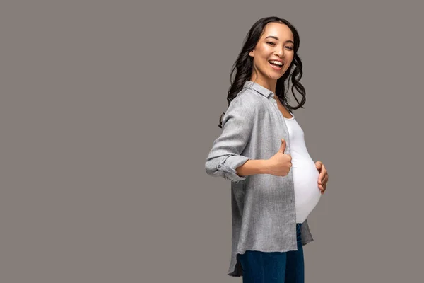 Sonriendo embarazada asiático mujer tocando vientre y mostrando pulgar arriba aislado en gris - foto de stock