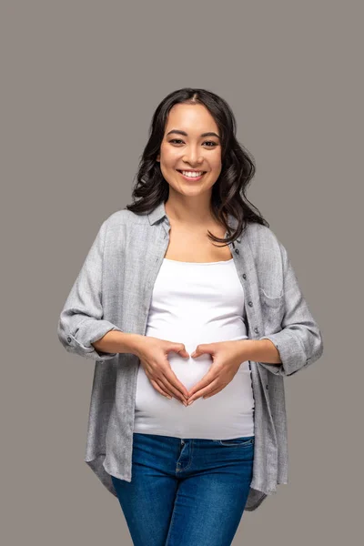 Молода усміхнена вагітна азіатка з руками показує серце на животі ізольовано на сірому — стокове фото