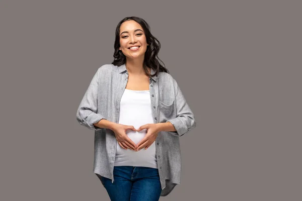 Молода щаслива вагітна азіатка з серцеподібними руками на животі ізольована на сірому — стокове фото