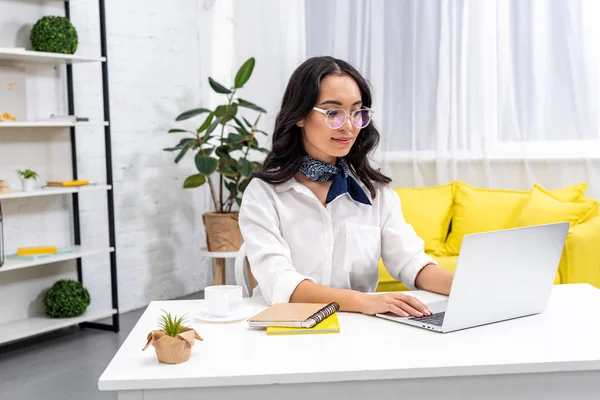 Молодий усміхнений азіатський фрілансер в окулярах, використовуючи ноутбук в домашньому офісі — стокове фото