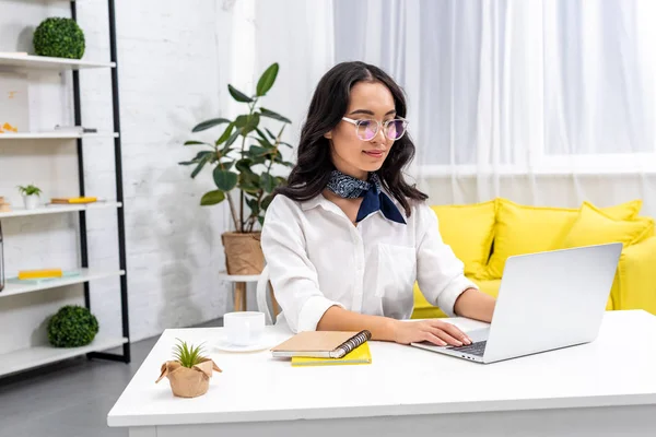 Jeune sourire asiatique pigiste dans des lunettes à l'aide d'un ordinateur portable au bureau à la maison — Photo de stock