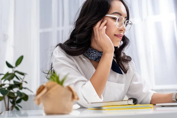 Selective focus of pretty asian freelancer in glasses sitting at work desk and holding hand on cheek — Stock Photo