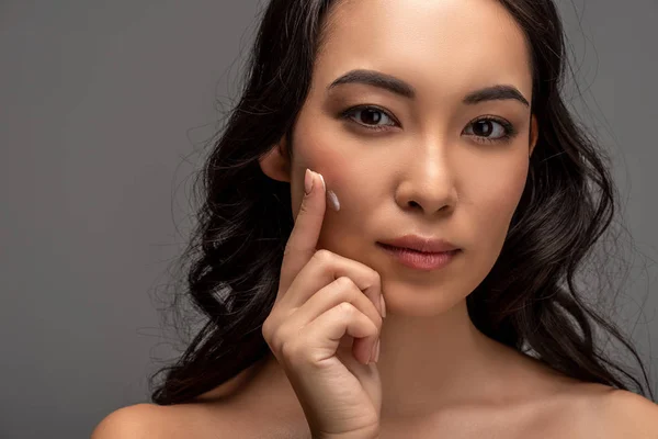 Portrait of beautiful asian girl applying cosmetic cream on face and looking at camera isolated on grey — Stock Photo