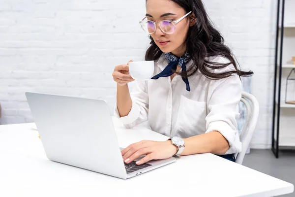 Young asian freelancer in glasses using laptop and drinking coffee at home workplace — Stock Photo