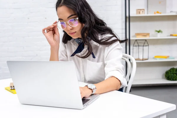 Thoughtful asian freelancer using laptop at home workplace and correcting glasses — Stock Photo