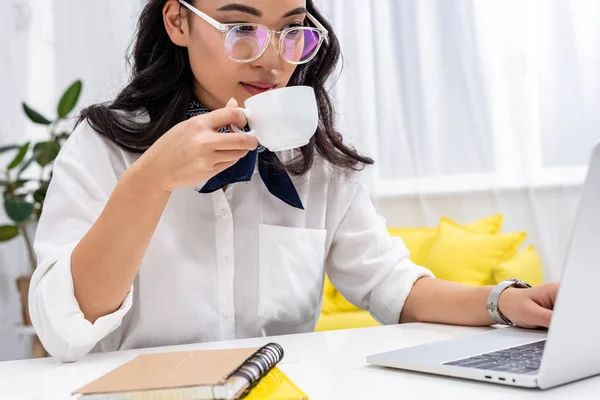 Attractive asian freelancer using laptop and drinking coffee at home workplace — Stock Photo