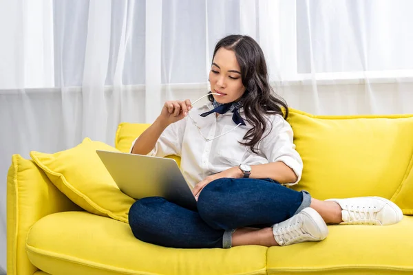 Young asian freelancer using laptop while sitting with feet on yellow sofa and holding glasses in hand — Stock Photo
