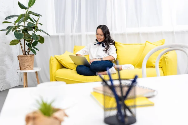 Foyer sélectif de pigiste asiatique assis avec les pieds sur le canapé jaune et en utilisant un ordinateur portable avec un lieu de travail flou au premier plan — Photo de stock